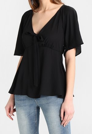 Kobieta ubrana w czarną bluzkę z dekoltem w serek, wiązaną z przodu i z falbanowatymi rękawami, w połączeniu z jasnoniebieskimi jeansami.