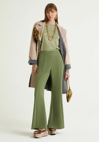 Top verde con motivo abbinato a pantaloni olivastro a zampa a vita alta, un cappotto beige e grigio e sandali con plateau. Accessoriato con una collana di perline.