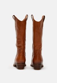 Botas de vaquero de cuero marrón con un caña alta, línea superior puntiaguda y tacón de madera apilada. Textura suave y detalles de costura mínimos.