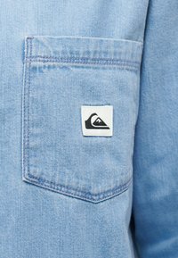 Κοντινό πλάνο ανοιχτό μπλε τζιν πουκάμισο που δείχνει μια τσέπη στο στήθος με ένα μικρό λευκό έμβλημα με μαύρο στυλιζαρισμένο κύμα και βουνό.