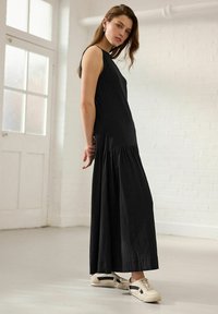 Rochie maxi fără mâneci, de culoare neagră, cu fustă plisată, realizată dintr-un material moale. Purtați-o cu adidași bej cu accente negre și textură netedă.