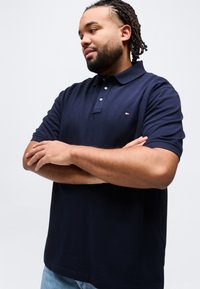 Άνδρας με κοτσιδωτά μαλλιά και γένια, φορώντας μπλε σκούρο πόλο και ανοιχτόχρωμο μπλε τζιν, στέκεται με σταυρωμένα τα χέρια μπροστά σε λευκό φόντο.