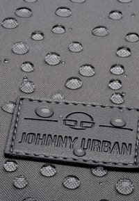 Tissu noir résistant à l'eau avec une texture à pois en relief, doté d'une étiquette en cuir embossée avec "JOHNNY URBAN" et de gouttes d'eau dispersées.