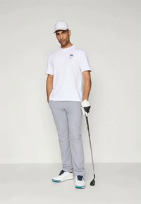 Γκολφ ενδυμασία που περιλαμβάνει ένα λευκό μπλουζάκι, ανοιχτό γκρίζο παντελόνι και λευκά παπούτσια με μπλε λεπτομέρειες. Κρατώντας μια γκολφ ρακέτα, φορώντας ένα λευκό καπέλο και γάντι.