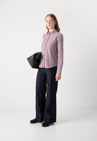 Camicia a righe rossa e bianca, pantaloni in denim scuri a gamba larga e borsa nera. Il modello indossa sneaker nere. Sfondo semplice.
