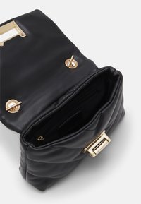 Černá prošívaná kožená kabelka otevřená, aby ukázala vnitřní zipovou kapsu a zlaté otočné uzávěry na bílém pozadí.