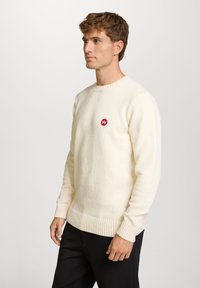Kremfarget strikket genser med tekstur, ribbestrikkede mansjetter og kant, med et lite rødt logo-patch på brystet.