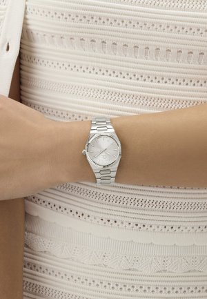 Sudraba rokas pulkstenis ar metāla siksniņu, nēsāts uz kreisās rokas plaukstas, uz fona balts teksturēts adīts tops.