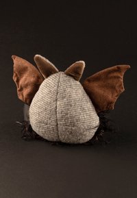 Gray and brown plush toy with pointed ears, bat-like wings, and a textured body. Base features dark, fluffy accents.