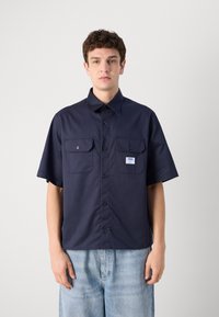 Mørkeblå kortermet skjorte med to framlommer, knappelukking og et lite logomerke, laget av et glatt stoff med en strukturert design.