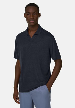 Junger Mann, der ein dunkelblaues Polohemd mit kurzen Ärmeln und hellblaue Hosen trägt und mit einer Hand in der Hosentasche vor einem einfarbigen Hintergrund steht.