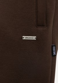 Nahaufnahme eines braunen Stoffes mit zwei SMILODOX-Markenlabels, einem mittig platzierten Metallplättchen und einem schwarzen Stoffetikett an der Seitennaht.