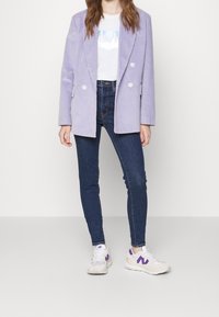 Mujer vestida con un blazer lavanda de doble botonadura, una camiseta gráfica blanca, unos jeans skinny en azul oscuro y unas zapatillas blancas con detalles morados.