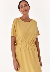 Gelbes Kurzarmkleid aus weichem Material mit lässiger Passform, das einen elastischen Bund und eine Zugbandschleife an der Seite aufweist.