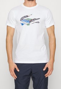White cotton t-shirt featuring a multicolored graphic of a crocodile logo composed of text in various fonts and colors. Short sleeves, round neckline.