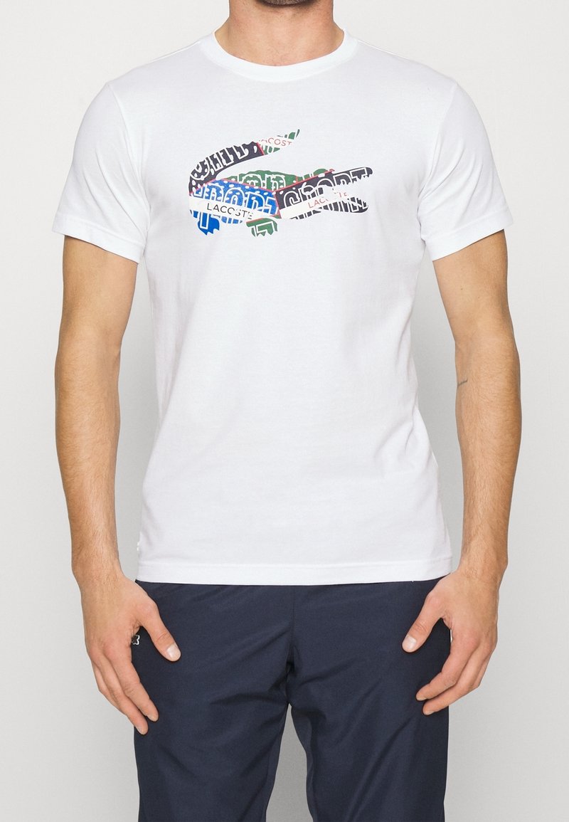 White cotton t-shirt featuring a multicolored graphic of a crocodile logo composed of text in various fonts and colors. Short sleeves, round neckline.