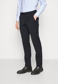 Pantaloni navy su misura realizzati in tessuto liscio, con tasche frontali e taglio dritto, abbinati a mocassini neri.