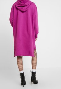 Person som bär en lång lila huvtröja, svarta spetstights, vita strumpor och svarta snörade högklackade stövletter, stående.