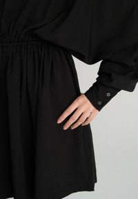 Czarna sukienka z marszczoną talią, długimi rękawami i zapięciem na guziki. Materiał jest gładki, z luźnym krojem i zwiewną sylwetką.