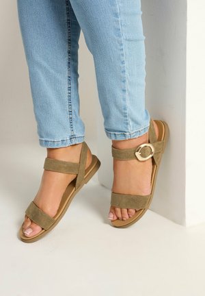 Jean bleu clair et sandales en daim beige avec une boucle dorée portés par une personne debout contre un mur blanc.