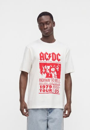 Jeune homme portant un t-shirt blanc du groupe AC/DC avec une impression rouge de la tournée Highway to Hell de 1979 et un jean bleu clair.