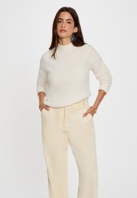 Crème-kleurige, textuurtrui met een hoge hals, gecombineerd met lichtbeige, geribbelde wijde broek. Opvallende chunky gouden sieraden als accent.