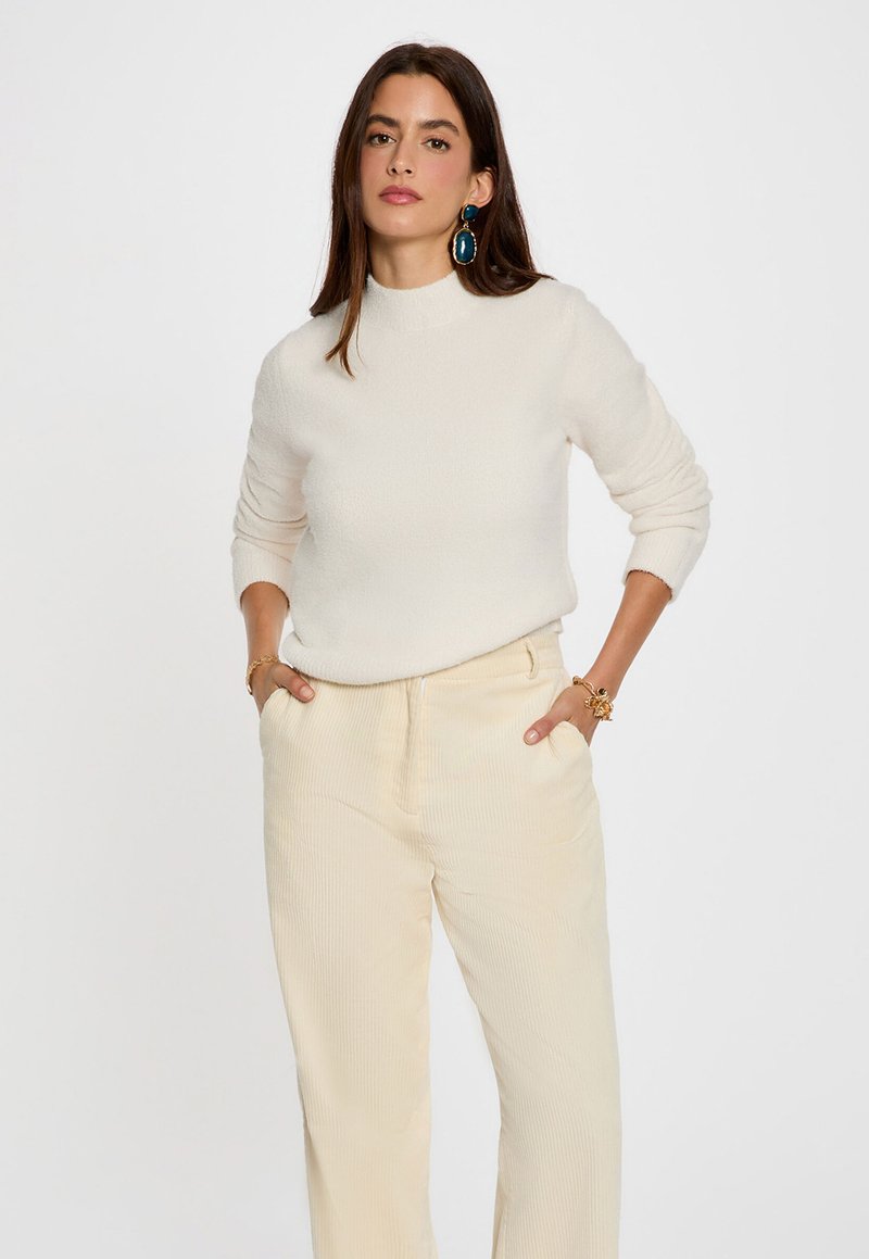 Crème-kleurige, textuurtrui met een hoge hals, gecombineerd met lichtbeige, geribbelde wijde broek. Opvallende chunky gouden sieraden als accent.