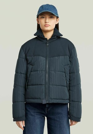 Junge Person trägt eine dunkelblaue, gestreifte Steppjacke mit Fleece-Kragen und eine blaue Jeans-Baseballkappe, steht vor einem neutralen Hintergrund.