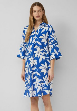 Femme portant une robe bleue mi-longue aux genoux avec de grands motifs floraux blancs, manches trois-quarts et col, debout devant un fond gris uni.