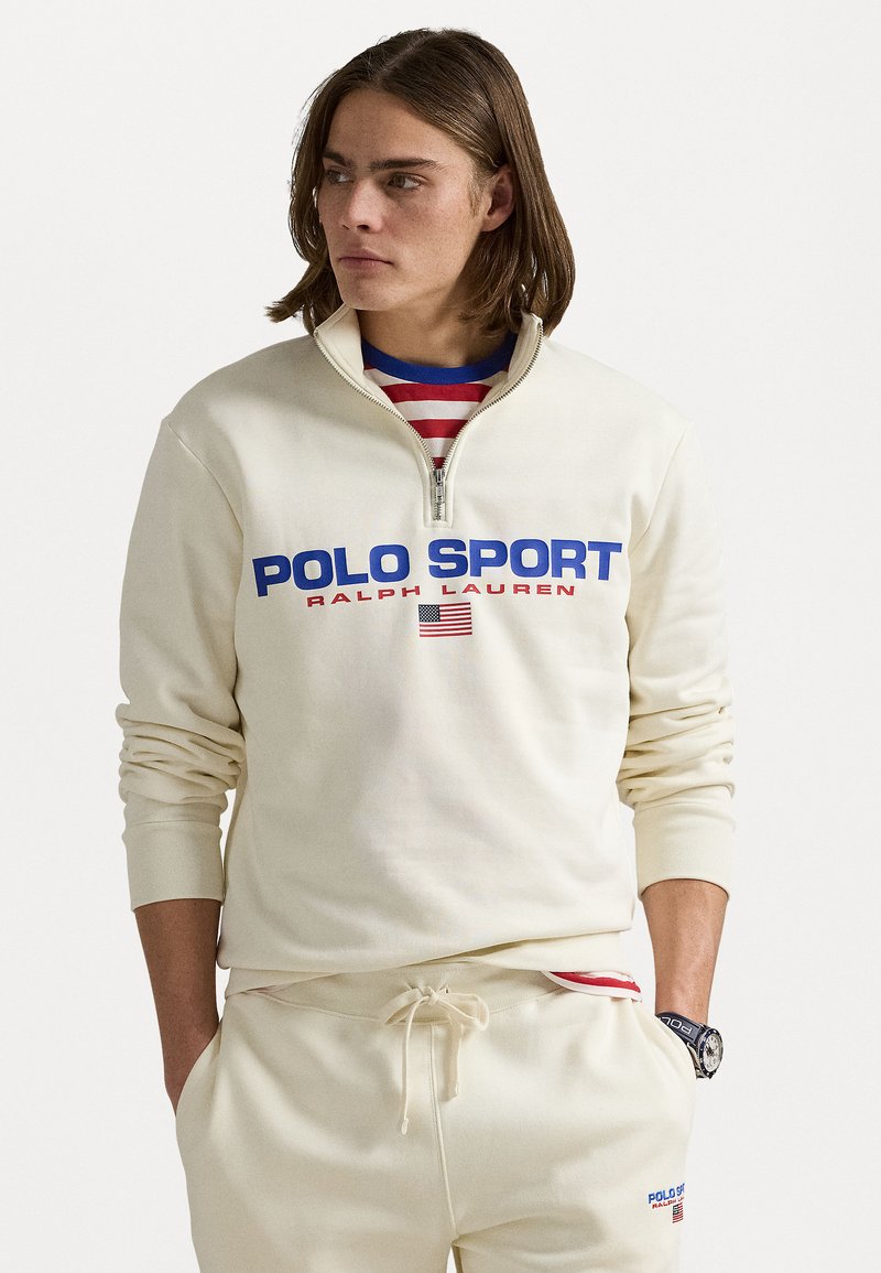 Hanorac crem cu fermoar, cu text și logo "POLO SPORT" în culorile albastru și roșu; manșete cu nervuri; purtat peste o cămașă cu dungi roșii și albe.