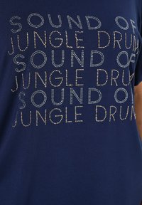 Námorně modré tričko s kovovými cvočky tvořícími text "SOUND OF JUNGLE DRUM" v různých velikostech a s různými mezerami přes celou přední stranu.