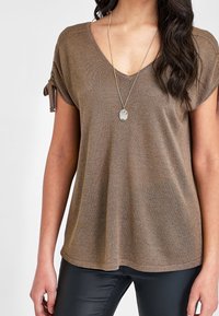 Mujer vistiendo una blusa de punto de manga corta marrón con detalles de mangas anudadas y un collar largo con un colgante ovalado de mármol.