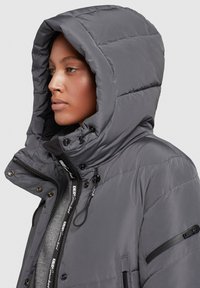 Graue gepolsterte Jacke mit großer Kapuze, Frontreißverschluss, bestickten Akzenten und seitlichen Reißverschlusstaschen. Glatte Textur mit gestepptem Design.