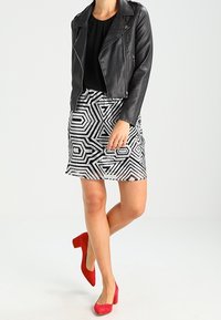 Mujer con chaqueta de cuero negra, blusa negra, falda de patrón geométrico y zapatos de tacón cuadrado rojos contra un fondo liso.