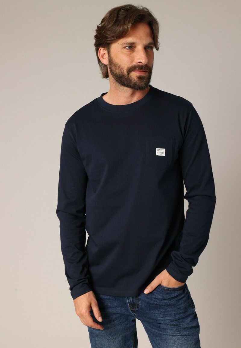 T-shirt à manches longues marine en coton, avec une poche poitrine dotée d'une étiquette. Le t-shirt a un col ras du cou et une coupe décontractée.