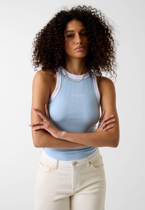 Vrouw met krullend haar draagt een lichtblauw mouwloos topje met witte rand en gebroken witte broek, staand met gekruiste armen tegen een effen achtergrond.