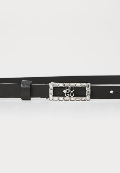 Cinturon noir en cuir mince avec une boucle rectangulaire en argent ornée de cristaux intégrés et du nom de la marque "HUGO" au centre.