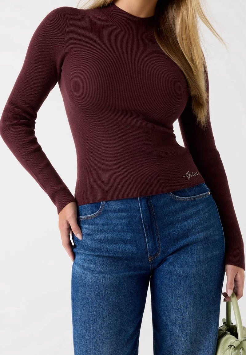 Femme portant un haut côtelé bordeaux à manches longues ajustées et un jean bleu, tenant un sac à main vert clair sur un fond uni.