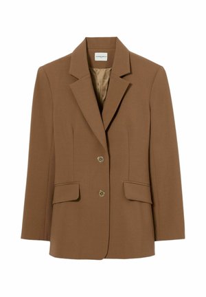 Mujer con un traje entallado color beige con pantalones de pierna ancha, una chaqueta abotonada y una pajarita marrón oscuro, de pie frente a un fondo blanco liso.