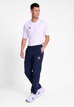 Weißes T-Shirt mit kurzen Ärmeln und Logo, kombiniert mit dunkelblauen Jogginghosen mit elastischem Bund und Chevron-Muster. Die Schuhe sind hell gefärbt.