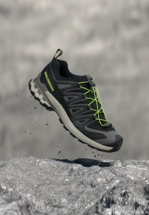 Zapatilla de trail running Salomon negra y gris con cordones verde neón que flota sobre un terreno rocoso con partículas de tierra en movimiento.