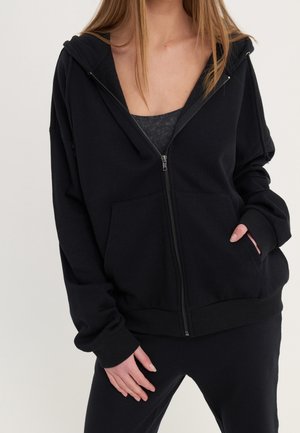 Personne portant un sweat à capuche noir zippé avec une poche avant sur un débardeur foncé et un pantalon noir assorti, mains détendues sur les côtés.