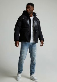 Svart dunjakke med blank finish, hette og frontlommer, kombinert med lyseblå revne jeans og hvite joggesko.