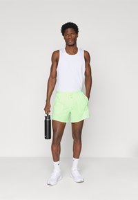 Νεόν πράσινα σορτς με κορδόνι, συνδυασμένα με λευκή φανέλα. Το άτομο κρατάει μια μαύρη φιάλη νερού, φοράει λευκά αθλητικά παπούτσια και κάλτσες.