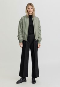 Groene bomberjack met ribgebreide manchetten en zoom, gecombineerd met wijde zwarte broek en zwarte schoenen. Minimalistisch ontwerp, gladde textuur.