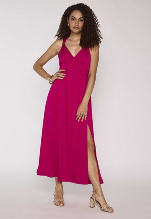 Donna con capelli ricci indossa un vestito senza maniche fucsia con spacco laterale e tacchi dorati con cinturino, accessoriata con anelli e braccialetti.
