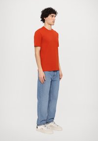 Red cotton T-shirt with short sleeves, featuring a subtle textured logo on the front. Worn with light blue jeans and sneakers.