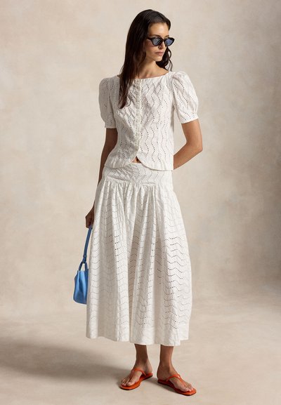 Γυναίκα που φοράει λευκό μπλουζάκι με μανίκια puff από eyelet ύφασμα και ταιριαστή midi φούστα, μαύρα γυαλιά ηλίου, πορτοκαλί σανδάλια, κρατώντας μικρή μπλε τσάντα.