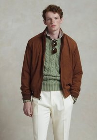 Blouson en daim marron sur un pull en tricot vert à texture, associé à un pantalon crémeux clair ; chemise à motif subtil et lunettes de soleil.