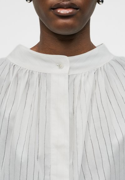 Κοντινό πλάνο ενός ατόμου που φοράει λευκό πουκάμισο με λεπτές κάθετες ρίγες και γιακά με ένα κουμπί, με εμφανή τα χείλη και το κάτω μέρος του προσώπου.